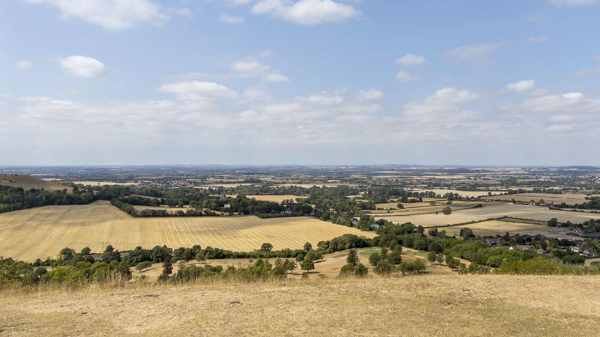 Panoramic view from Coombe Hill across rolling Chiltern farmland, with golden fields, scattered woodland and villages beneath a wide blue sky.