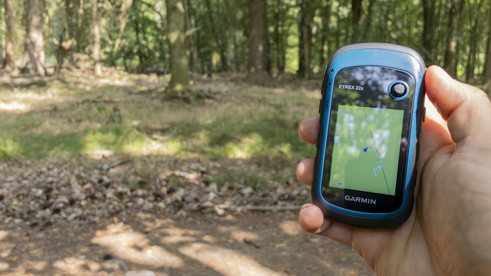 Hand holding a Garmin eTrex 22x GPS in woodland, with the map screen showing a small wandering track while the device remains stationary.