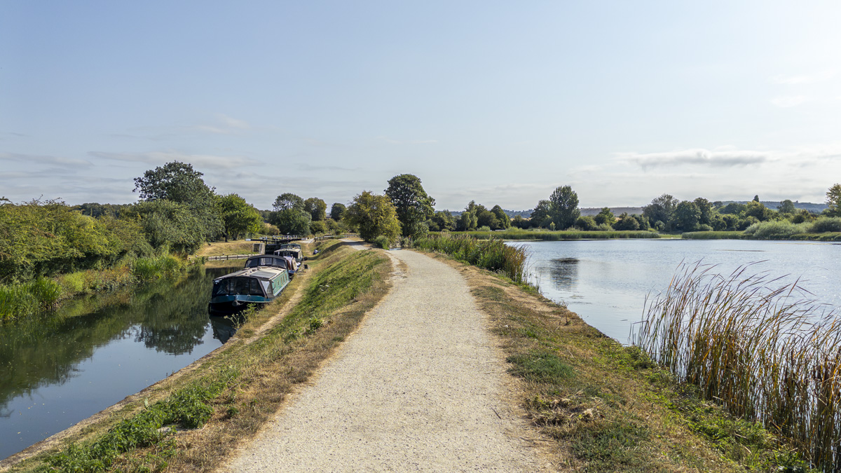 Gravel path between the Grand Union Canal, with moored narrowboats, and Marsworth Reservoir, bordered by reeds and trees under a clear blue sky.