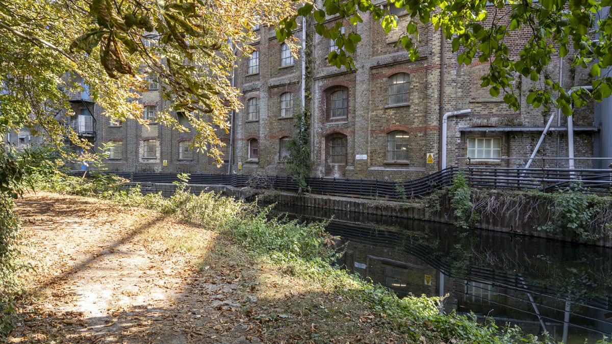 Historic brick mill beside the Grand Union Canal, with a shaded towpath running alongside the water beneath overhanging trees.