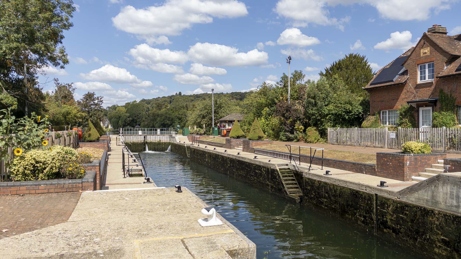 Hambledon Lock on the River Thames, with the lock chamber, mooring bollards and lockside cottage beneath a bright blue sky with scattered clouds.