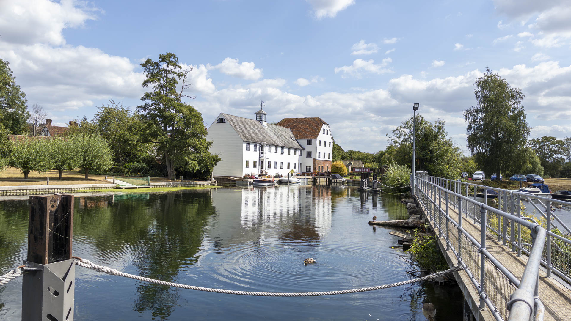 Hambledon Mill beside the River Thames, with the metal walkway crossing the water and trees lining the riverbank under a bright, partly cloudy sky.