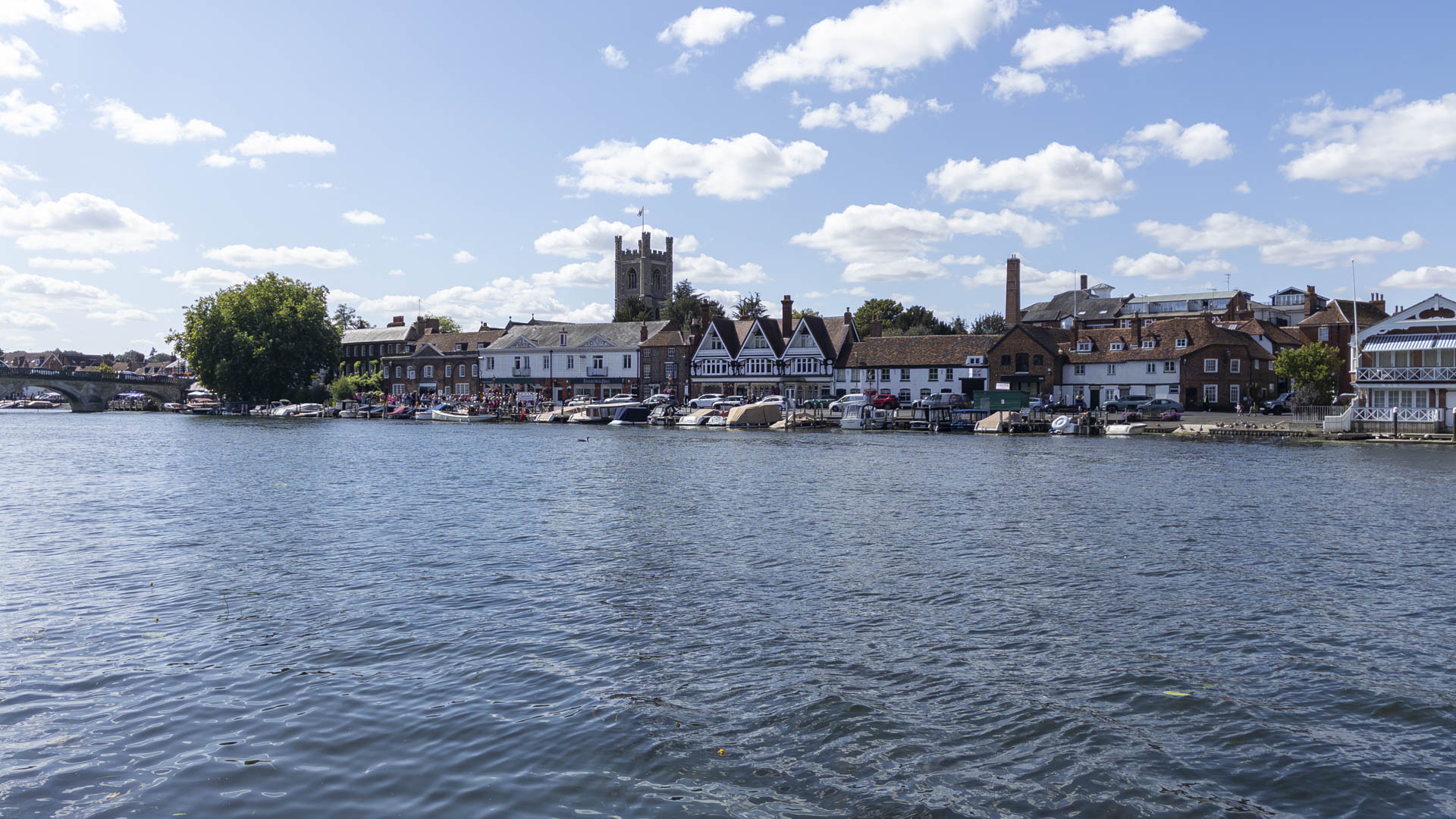 River Thames at Henley with boats moored along the waterfront, historic riverside buildings, a church tower and Henley Bridge beneath a bright blue sky.
